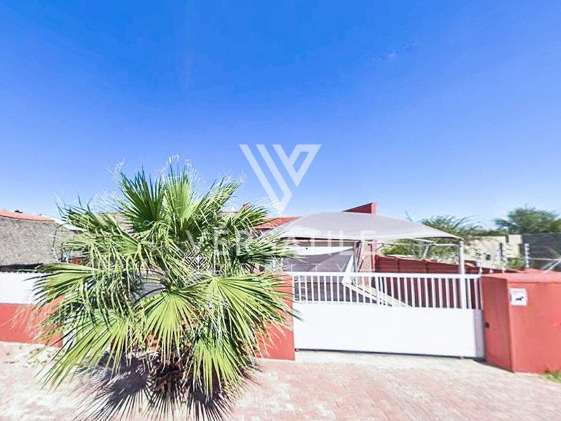Front exterior view of the property featuring a paved driveway, secure perimeter wall with electric fencing, and a shaded parking area.