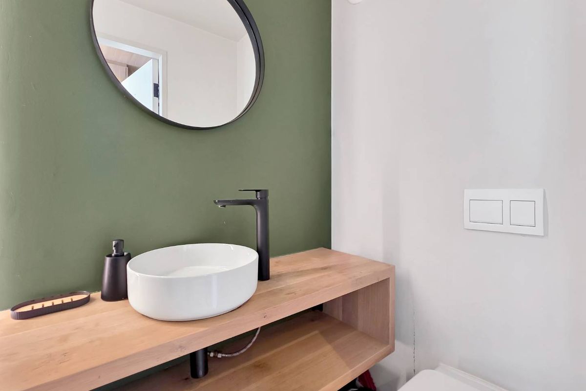 Modern bathroom featuring a timber vanity, white vessel sink with black tapware, and a circular mirror against a green accent wall.