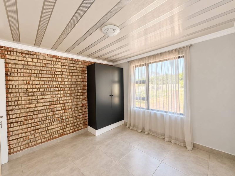 A bright bedroom featuring an exposed brick wall, tiled flooring, modern ceiling paneling, and a built-in cupboard with large window views.