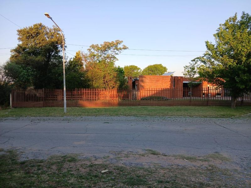 Street view of a single-story brick house featuring metal perimeter fencing, mature trees, and solar panels on the roof.