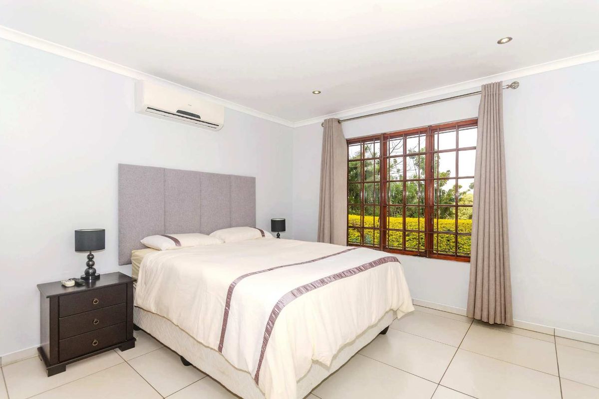 A bright bedroom featuring tiled flooring, a wall-mounted air conditioning unit, and large windows with garden views.