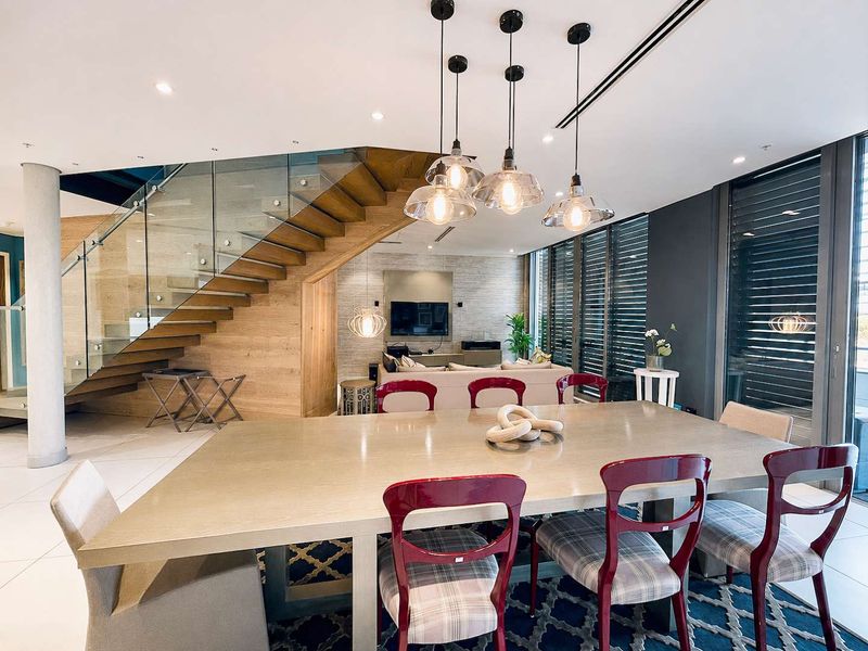 Modern open-plan living and dining area featuring a striking floating staircase with glass balustrade, high-end finishes, and abundant natural light through large sliding doors.