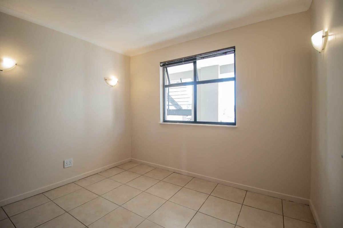 A bright, empty bedroom featuring neutral tiled flooring, wall-mounted lighting, and a large window providing natural light.