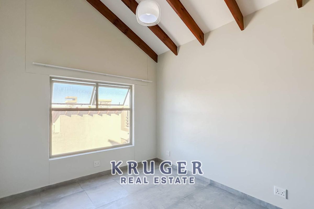A bright, empty bedroom featuring high vaulted ceilings with exposed timber beams, tiled flooring, and a large window providing natural light.