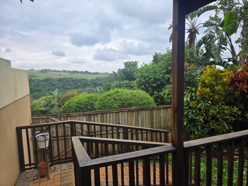 A covered patio area featuring a wooden balustrade and paved flooring, offering scenic views of the surrounding landscape and lush garden.