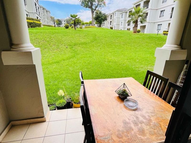 A covered patio area featuring tiled flooring and structural columns, overlooking a well-maintained communal garden and apartment complex grounds.