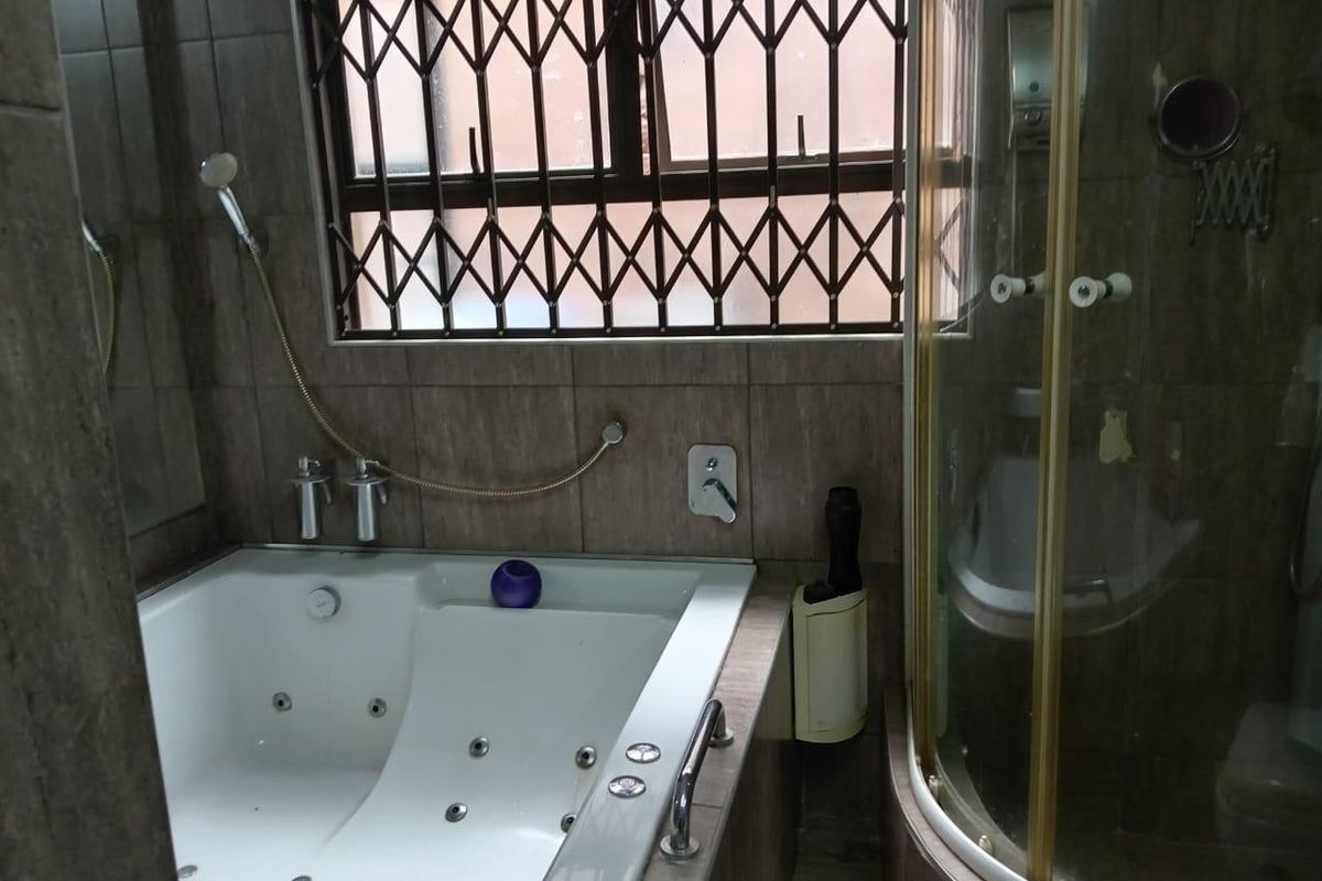 A functional bathroom featuring a built-in spa bath, a corner shower enclosure, and tiled finishes, complete with a security gate on the window.