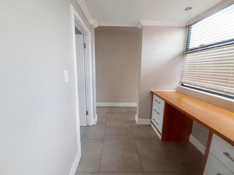 A bright, empty home office featuring a built-in wooden desk with integrated storage drawers, tiled flooring, and large windows with fitted blinds.