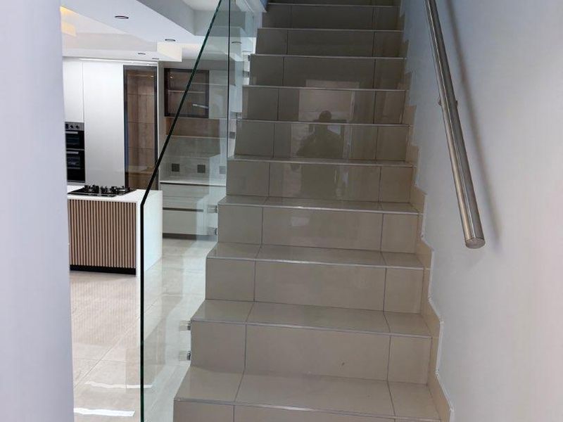 A modern hallway featuring a tiled staircase with a sleek glass balustrade and stainless steel handrail, leading towards an open-plan kitchen area.