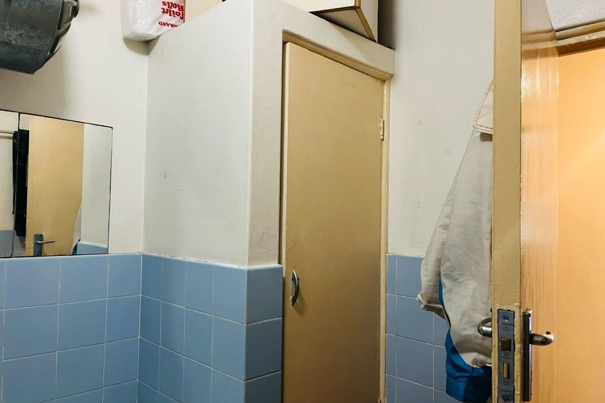 A functional bathroom space featuring blue wall tiles, a built-in storage cupboard, and a wall-mounted mirror, offering potential for modern cosmetic updates.