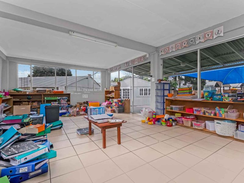 A spacious, light-filled room featuring tiled flooring and large windows, currently utilized as a classroom or play area with extensive storage shelving.