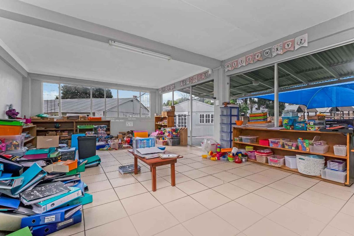 A spacious, light-filled room featuring tiled flooring and large windows, currently utilized as a classroom or play area with extensive storage shelving.