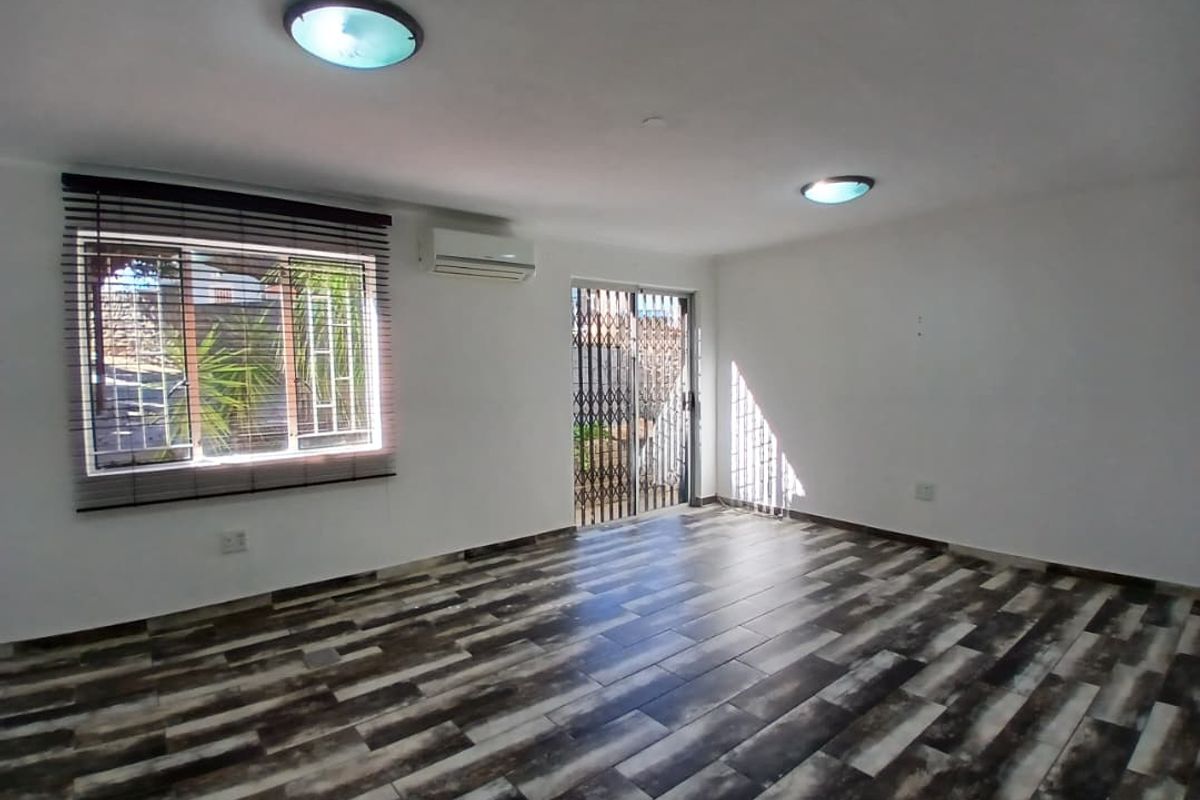 A bright, empty living room featuring modern wood-look tiled flooring, a wall-mounted air conditioning unit, and security gates on the sliding door.