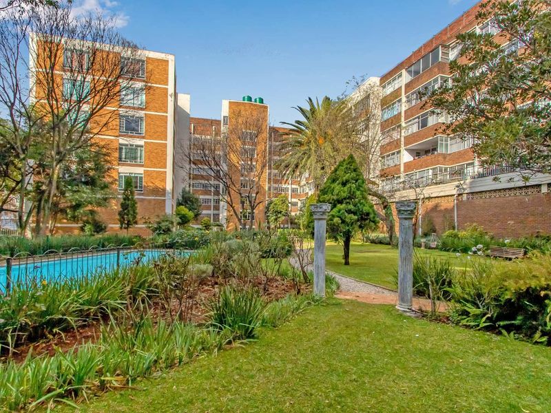 A view of the apartment complex exterior featuring a well-maintained communal garden, swimming pool, and decorative stone pillars set against the brick building facade.