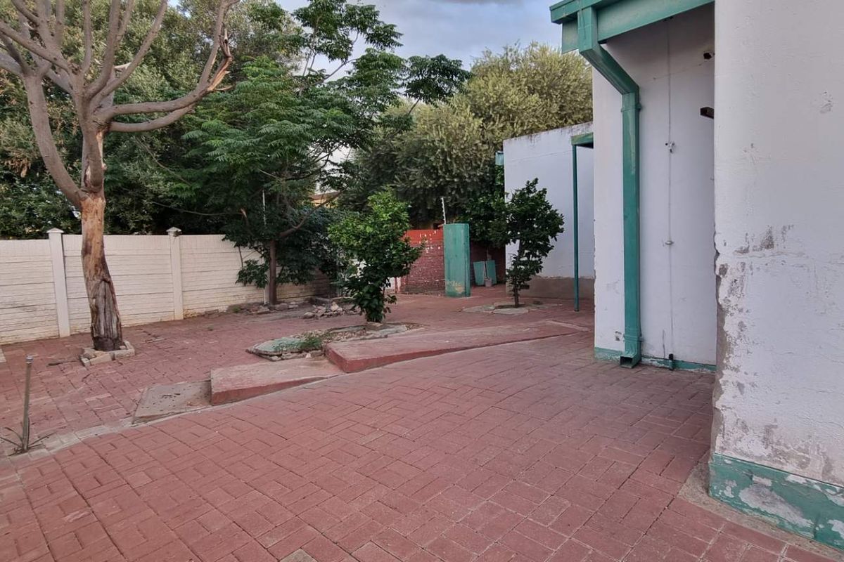 A rear exterior view of the property featuring a paved courtyard area, perimeter boundary walls, and established trees.