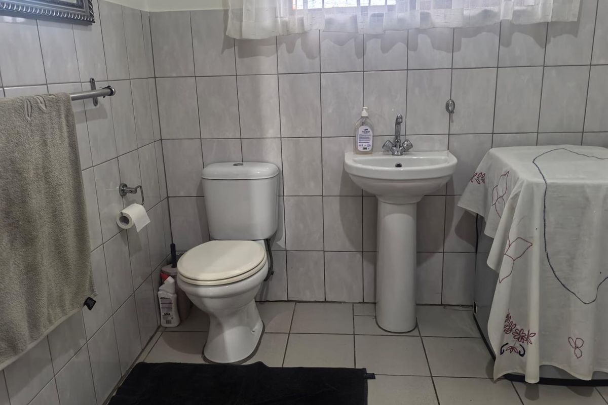 A functional bathroom featuring a pedestal washbasin, ceramic toilet, and full-height wall tiling with security bars on the window.