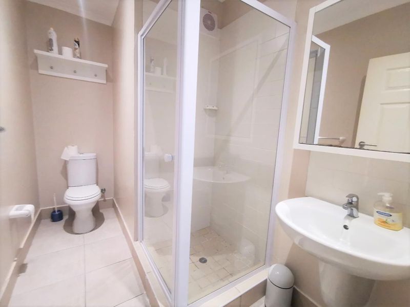 A clean and functional bathroom featuring a glass shower enclosure, pedestal basin, toilet, and wall-mounted mirror with neutral tiled flooring.