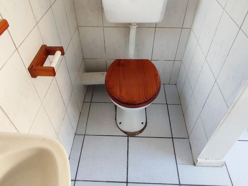 A compact bathroom featuring white ceramic wall and floor tiles, a standard toilet with a wooden seat, and a small wall-mounted washbasin.