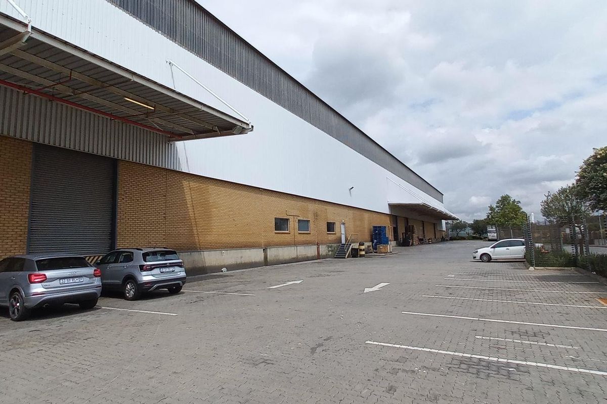 Industrial warehouse exterior featuring a large brick facade, roller shutter loading doors, and a paved parking area with perimeter security fencing.
