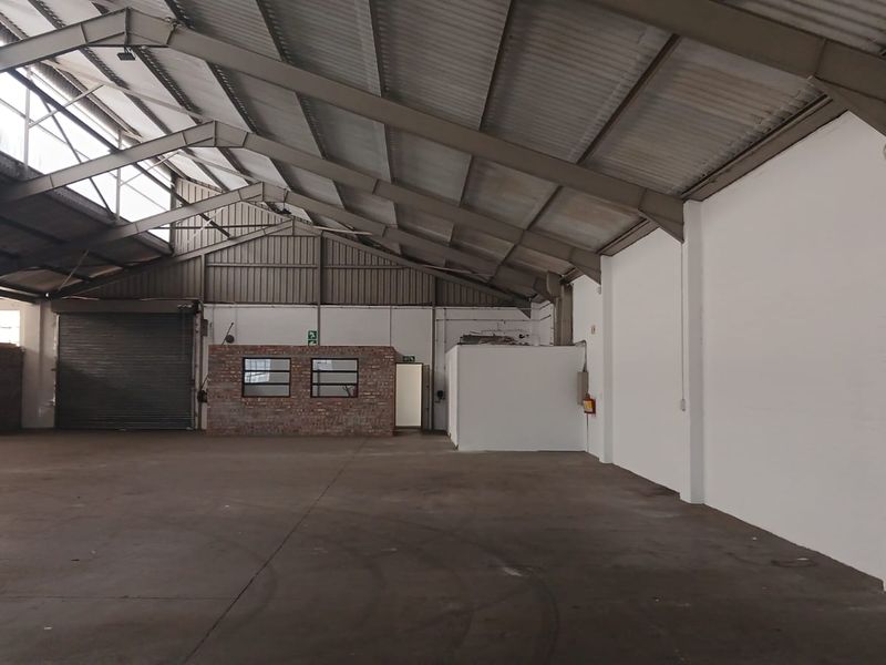 Spacious industrial warehouse unit featuring high ceilings with exposed steel trusses, concrete flooring, and a large roller shutter door for easy access.