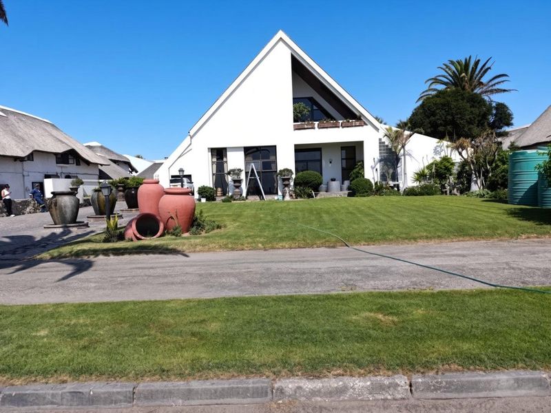 A well-maintained modern house with a distinctive pitched roof, balcony, and landscaped front lawn, featuring a paved driveway and exterior water storage.