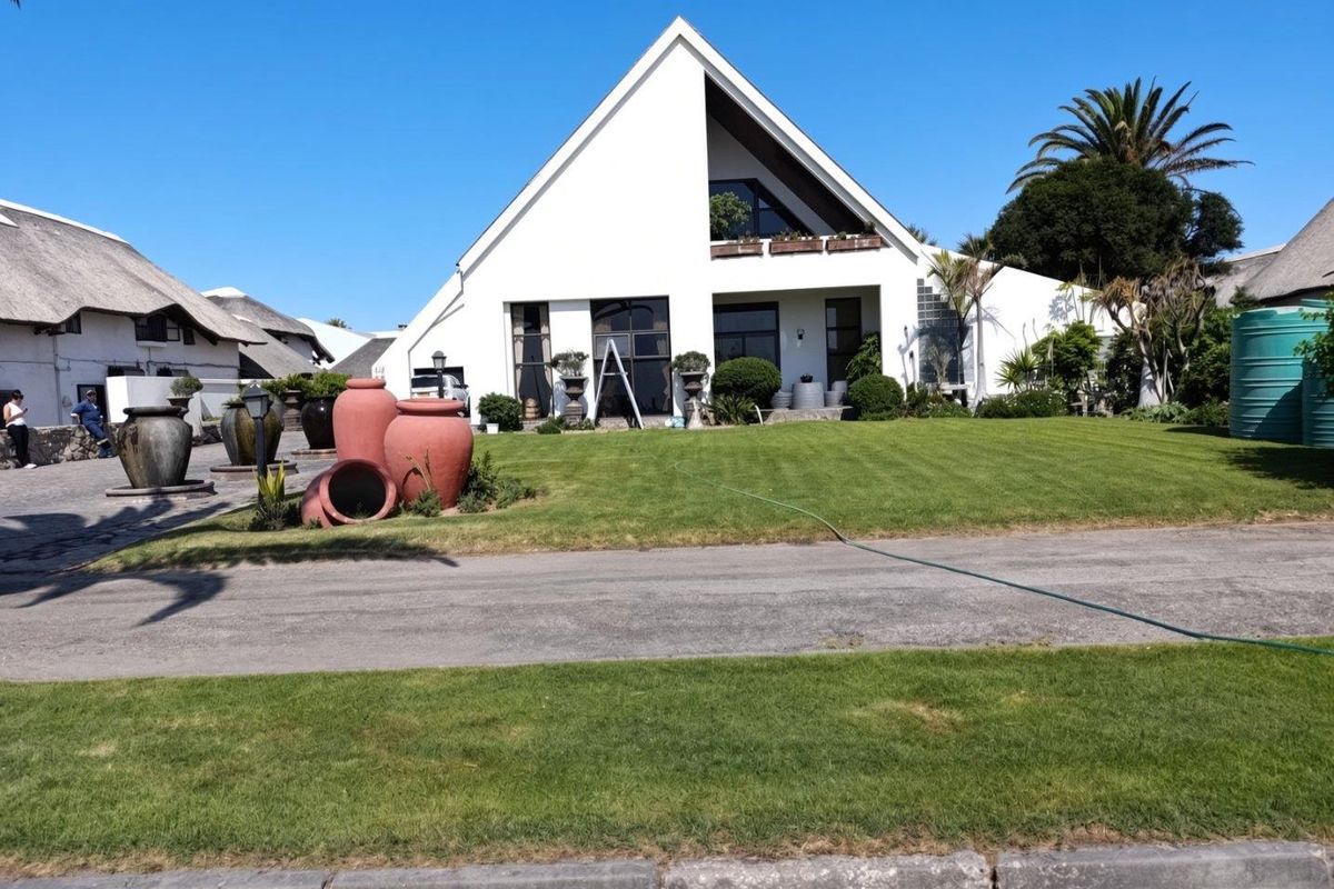 A well-maintained modern house with a distinctive pitched roof, balcony, and landscaped front lawn, featuring a paved driveway and exterior water storage.