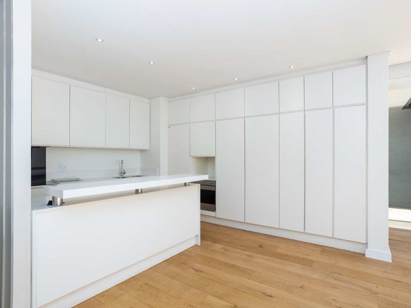 Modern minimalist kitchen featuring sleek white cabinetry, integrated oven, a functional breakfast bar island, and warm timber flooring.