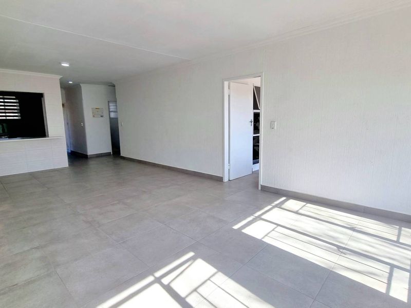 A spacious, empty open-plan living area featuring neutral tiled flooring, clean white walls, and a convenient pass-through window connecting to the kitchen.