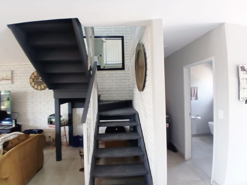 Open plan living area featuring a modern dark staircase, exposed white brick feature wall, and neutral tiled flooring leading to a secondary room.