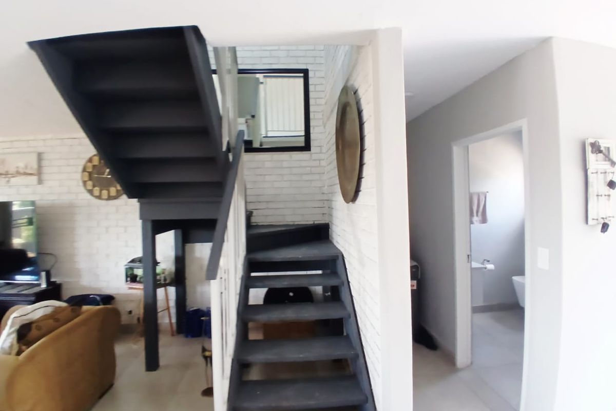Open plan living area featuring a modern dark staircase, exposed white brick feature wall, and neutral tiled flooring leading to a secondary room.