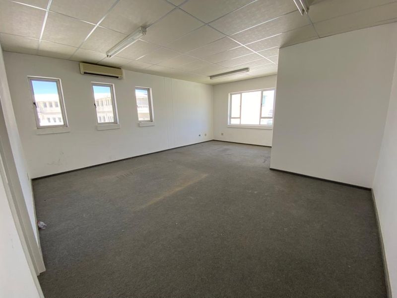 An empty office space featuring a suspended ceiling, wall-mounted air conditioning unit, and carpet flooring with multiple windows providing natural light.