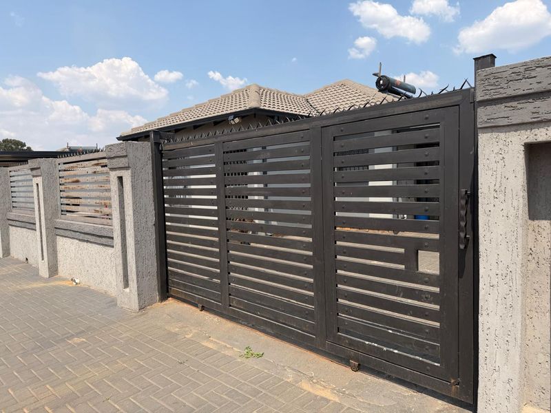 Front view of the property featuring a black metal sliding gate, perimeter wall with security spikes, and a paved driveway.