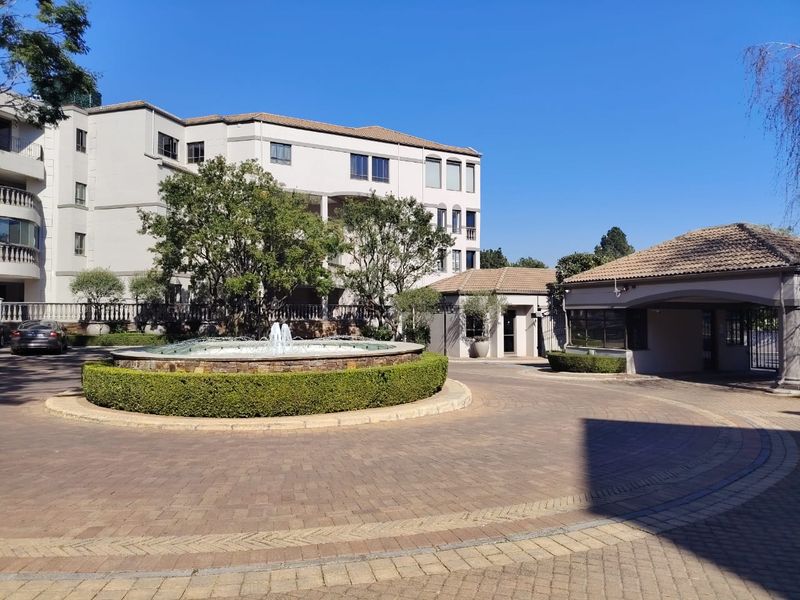 A well-maintained apartment complex entrance featuring a paved circular driveway, a central stone fountain, and a secure gatehouse.