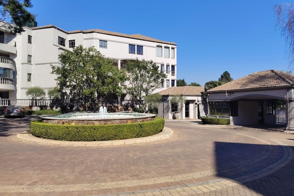 A well-maintained apartment complex entrance featuring a paved circular driveway, a central stone fountain, and a secure gatehouse.