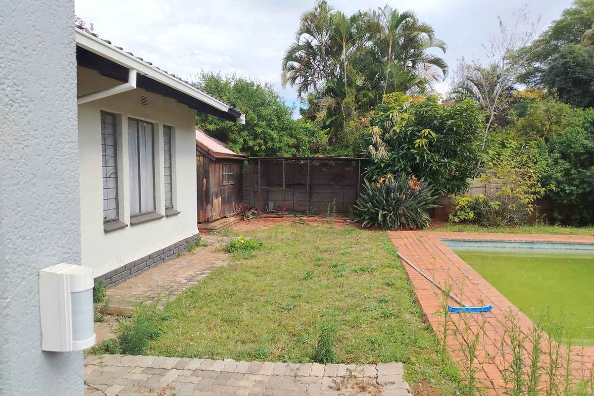 Rear garden area featuring a swimming pool, paved surround, and a weathered wooden outbuilding, set against a backdrop of mature trees.