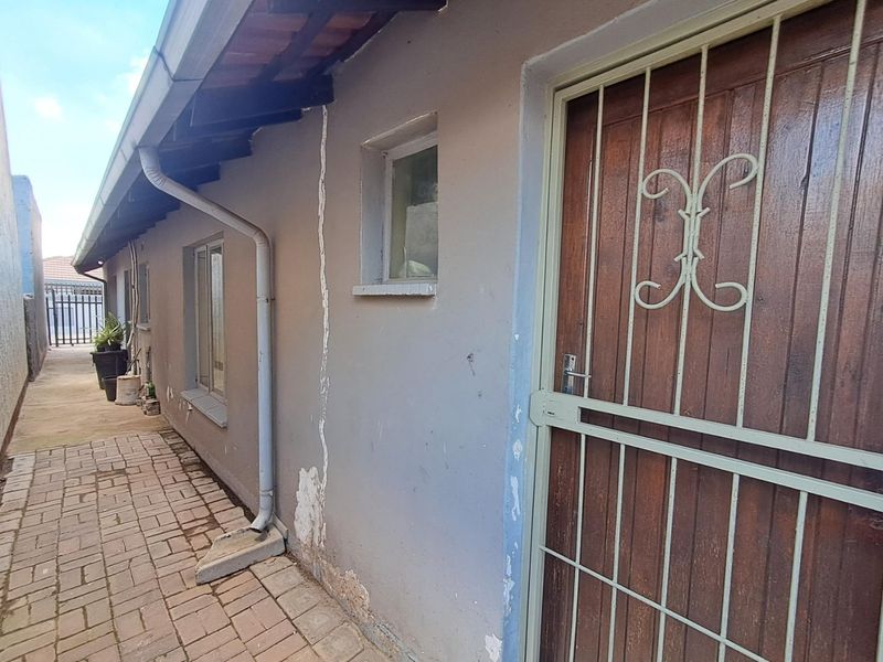 Side exterior view of the property featuring a paved walkway, timber door with security gate, and visible wall plaster damage.