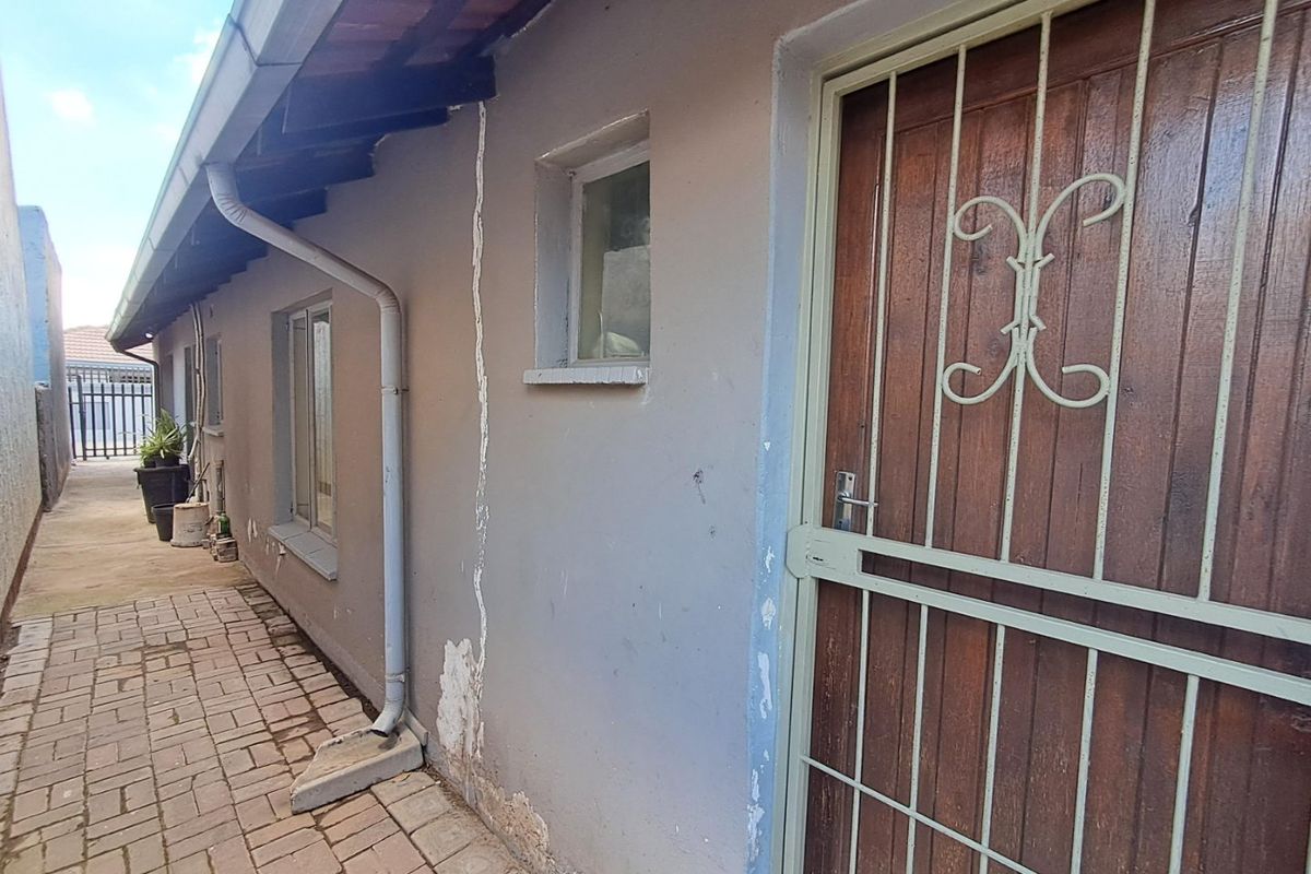 Side exterior view of the property featuring a paved walkway, timber door with security gate, and visible wall plaster damage.