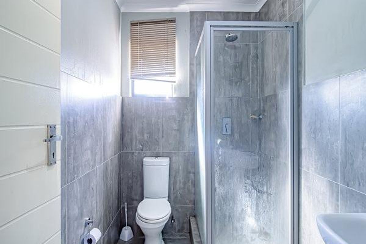 A compact, modern bathroom featuring grey wall and floor tiles, a glass shower enclosure, a white toilet, and a wall-mounted basin.