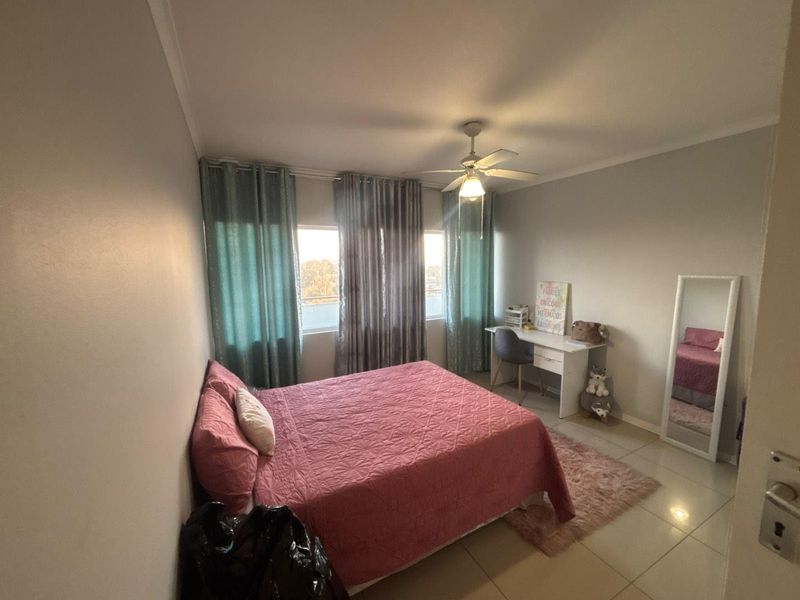 A bedroom featuring tiled flooring, a ceiling fan, and large windows with curtain tracks, providing a functional and well-lit living space.