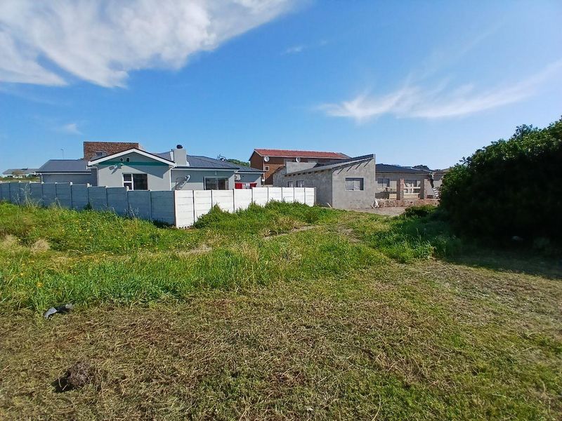 A view of a vacant residential plot featuring overgrown grass and a precast concrete boundary wall, adjacent to existing residential properties.