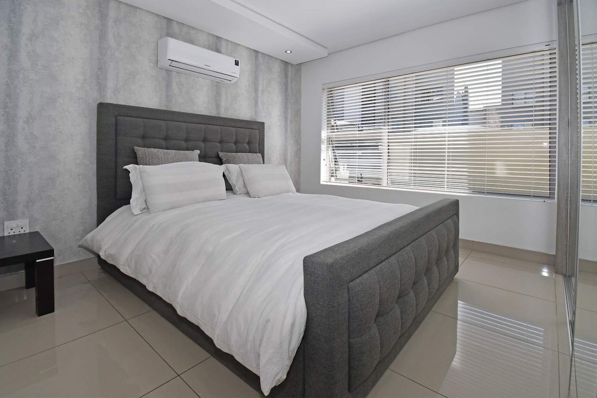Modern bedroom featuring neutral grey tones, polished porcelain floor tiles, a split-system air conditioner, and large windows with fitted venetian blinds.