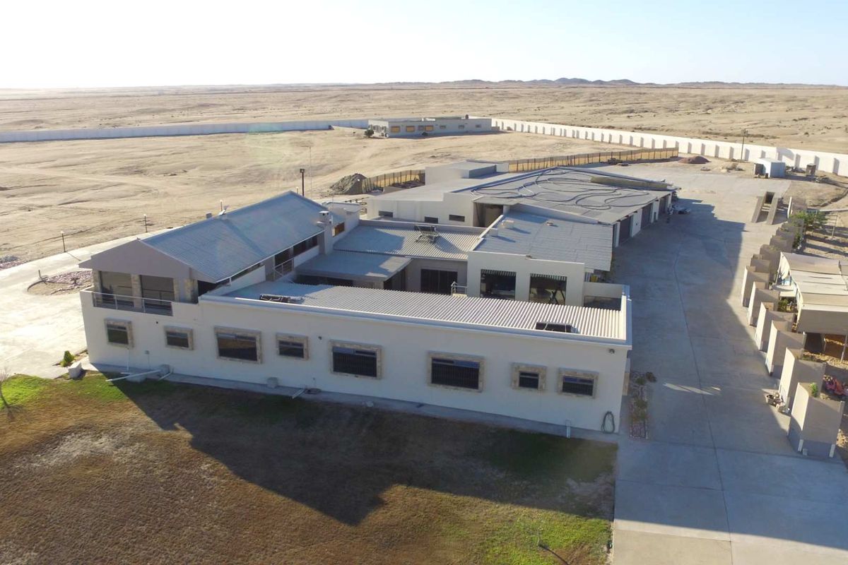 Aerial view of a large property featuring a multi-section building with metal roofing, extensive paved driveway areas, and surrounding desert landscape.