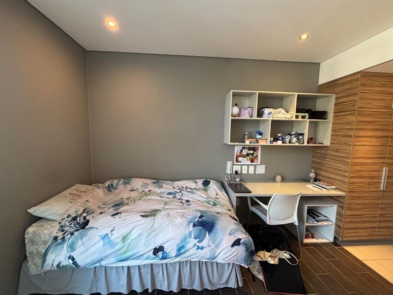 A functional bedroom featuring a built-in desk, integrated shelving unit, and wood-finish built-in wardrobe with recessed ceiling lighting.