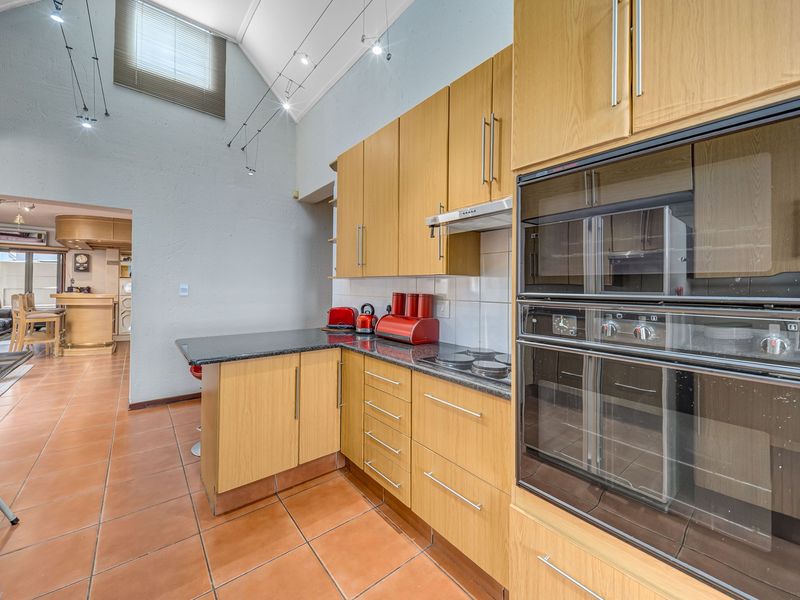 A functional kitchen featuring timber veneer cabinetry, granite countertops, built-in double oven, and electric hob, set under a high vaulted ceiling with tiled flooring.