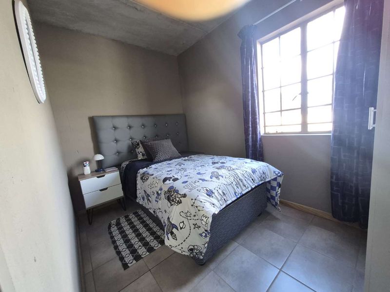 A compact bedroom featuring tiled flooring and a steel-framed window, with an unfinished concrete ceiling and plain wall surfaces.