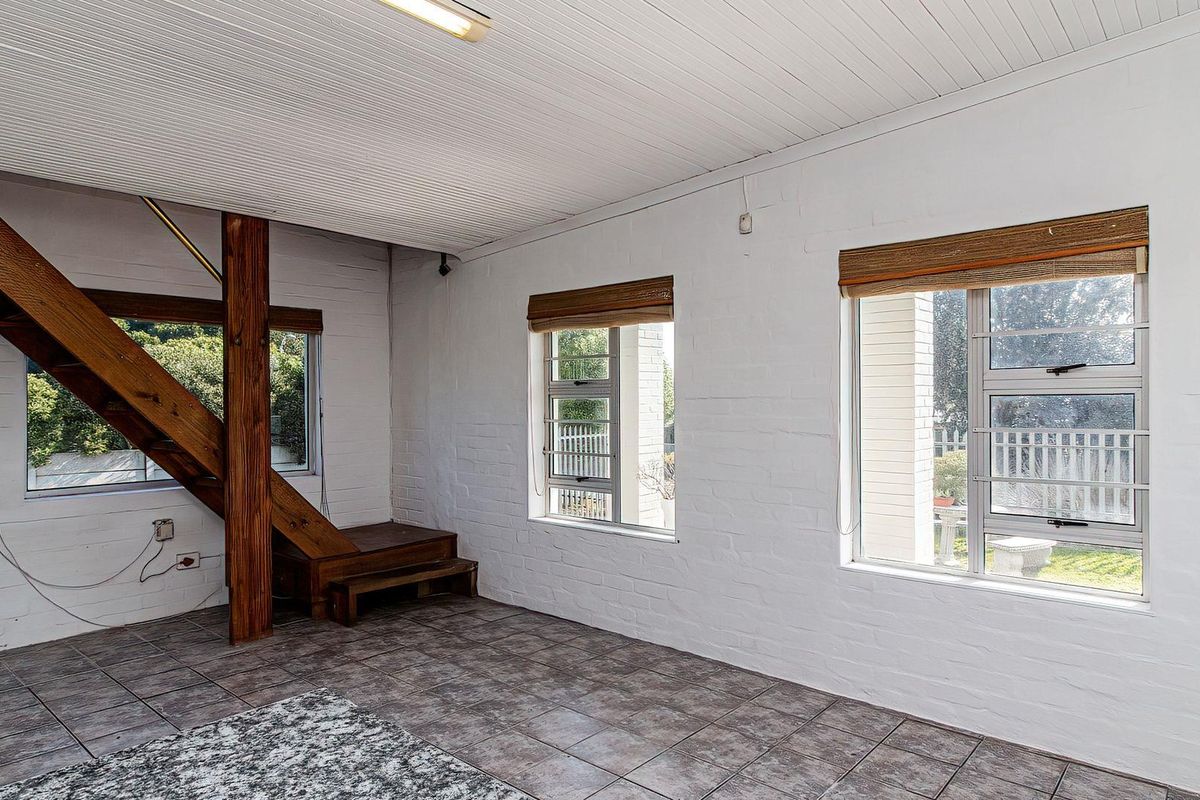 An open-plan living area featuring painted brick walls, tiled flooring, a timber staircase, and large windows providing natural light.