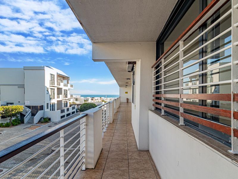A spacious, covered balcony featuring tiled flooring and a secure metal balustrade with wooden handrails, offering views of the surrounding apartment complex.