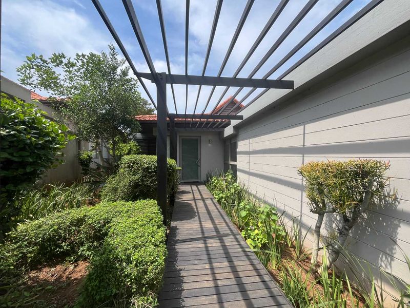 A well-maintained front entrance featuring a timber walkway, modern metal pergola, and established garden landscaping leading to the main door.