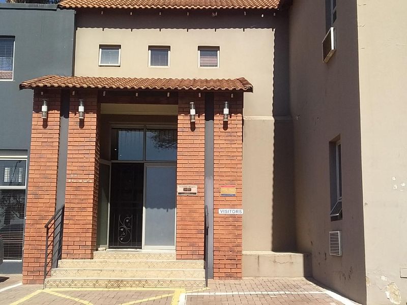Front entrance of a commercial building featuring a brick facade, tiled roof detail, and paved visitor parking area.
