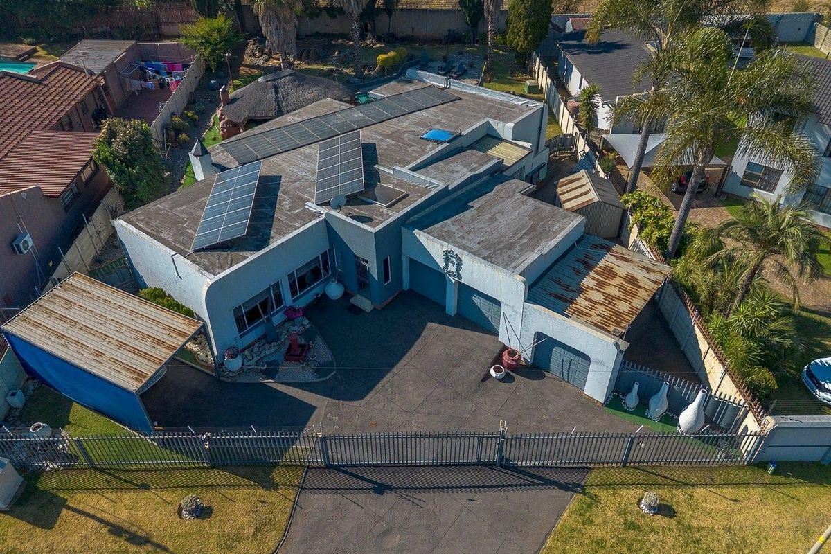 Aerial view of a residential property featuring a flat roof with solar panels, a double garage, asphalt driveway, and additional metal carport structures.